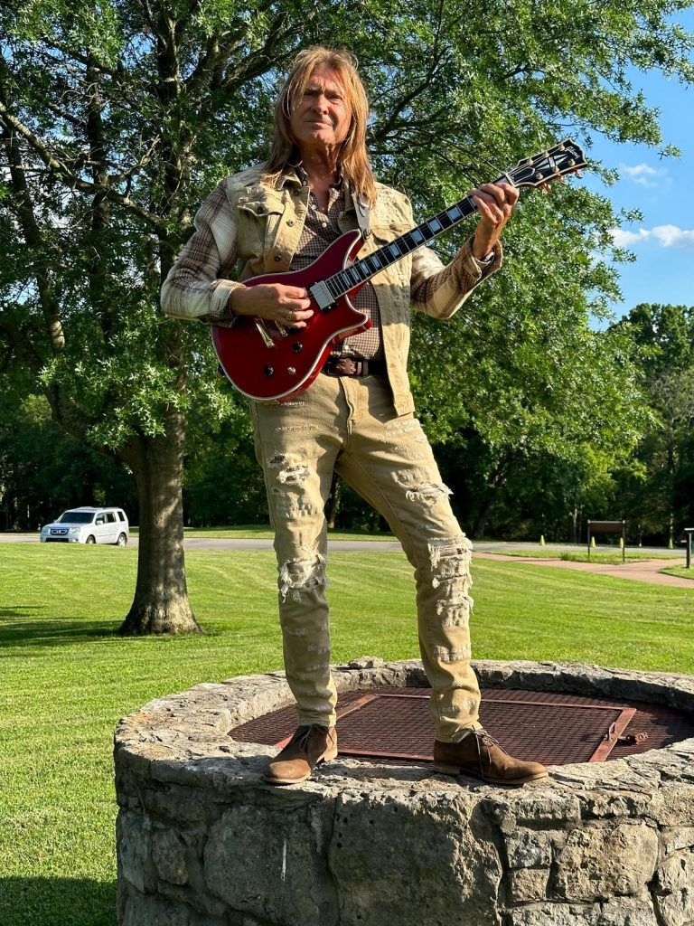 Man playing red guitar, standing on a stone structure outdoors. He wears a patterned jacket, ripped pants, and boots.