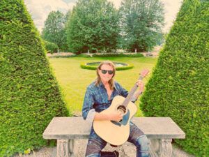 Man with sunglasses playing guitar on a stone bench in a formal garden with trees.