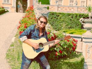 Man playing acoustic guitar outdoors in front of roses and a building.