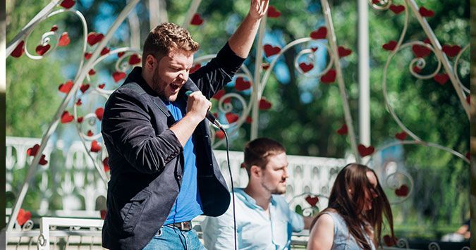 Man singing into a microphone, arm raised, standing on a stage with two others. Outdoor setting with heart decorations.