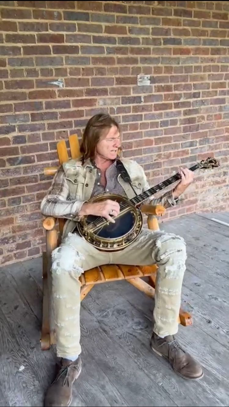 Man playing a banjo while sitting in a wooden rocking chair on a porch, brick background.