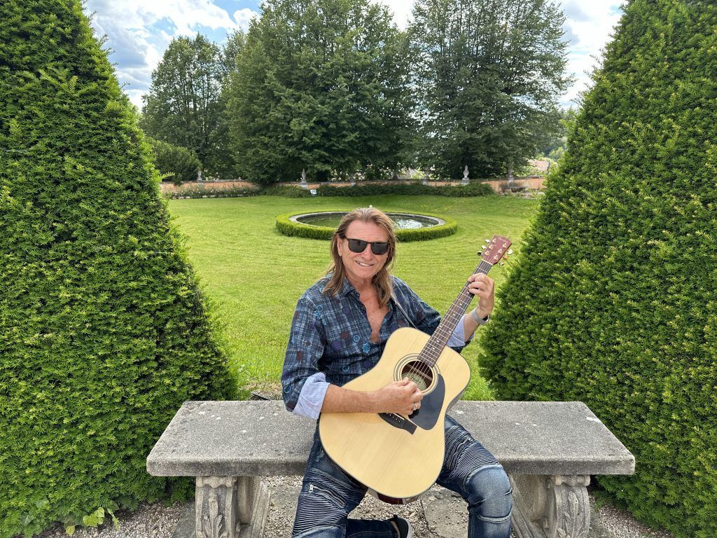 Man with long hair plays acoustic guitar outdoors on a stone bench.