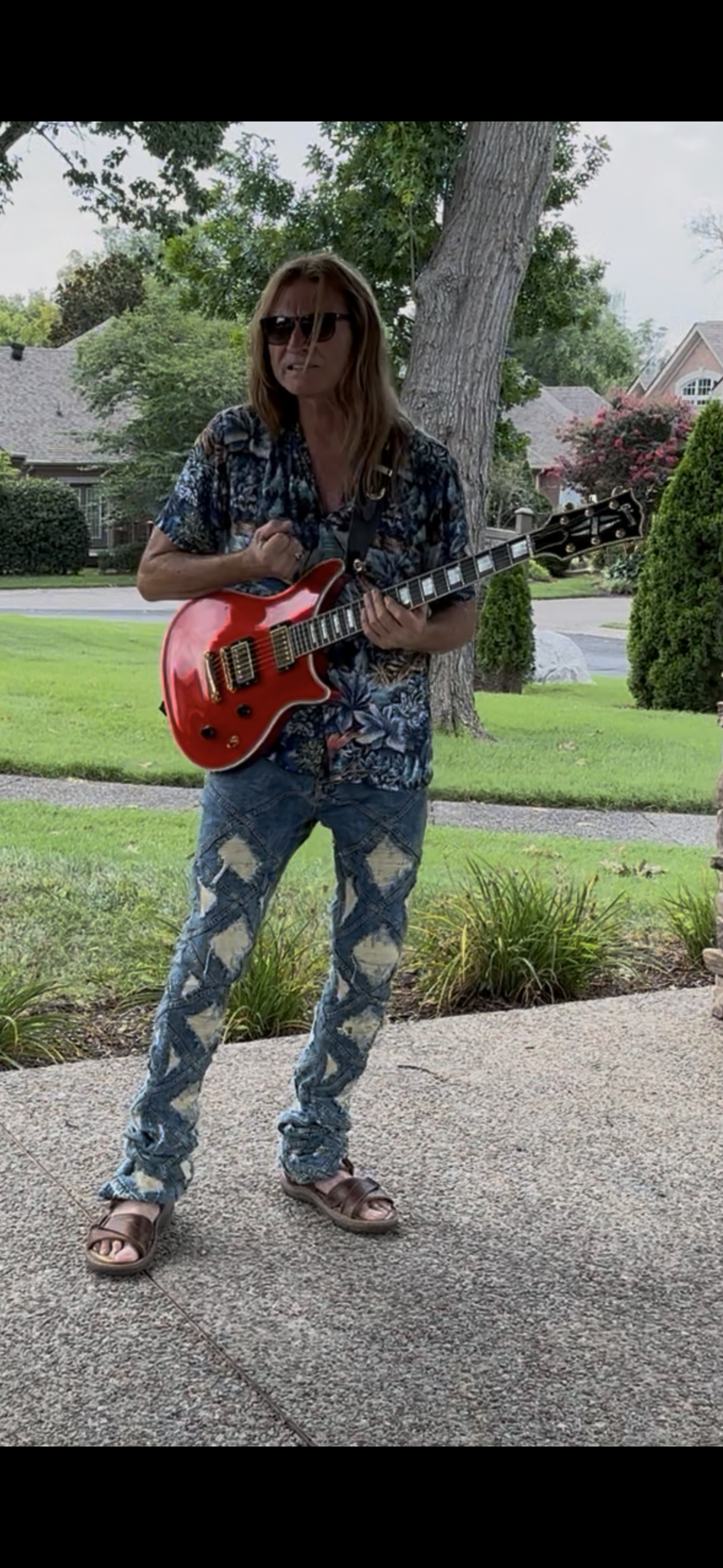 Person playing a red guitar outside, wearing sunglasses, patterned shirt, ripped jeans, and sandals.