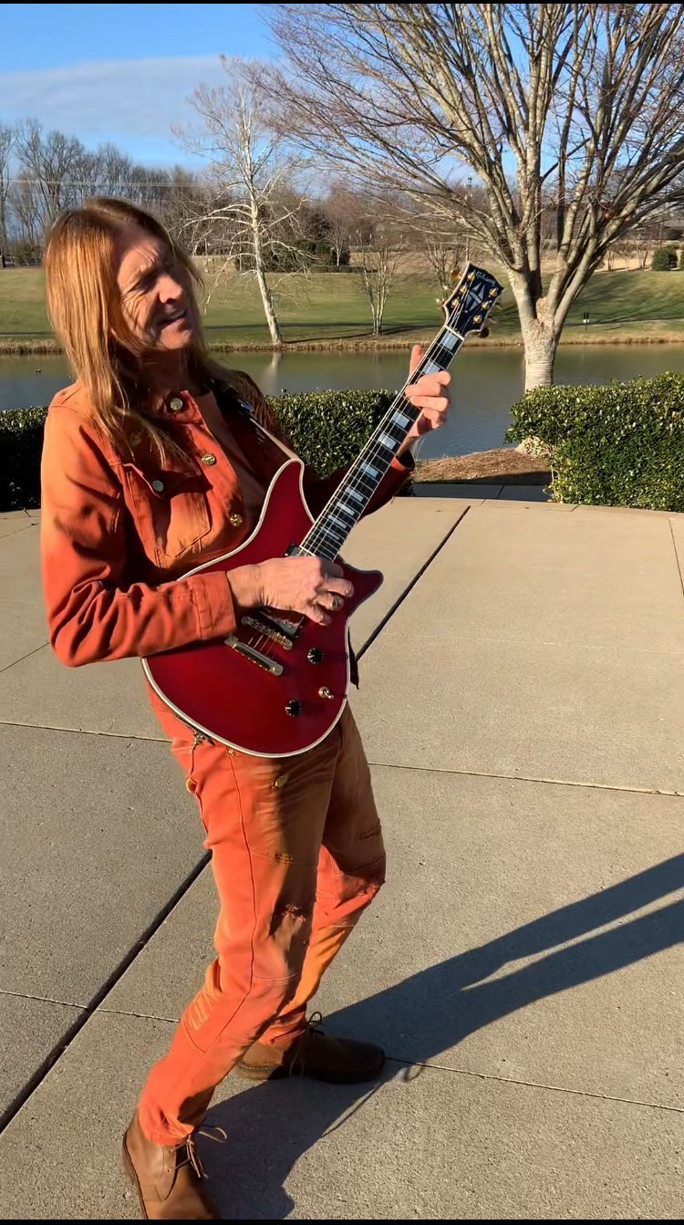 Person in orange outfit playing red electric guitar outside by a lake.