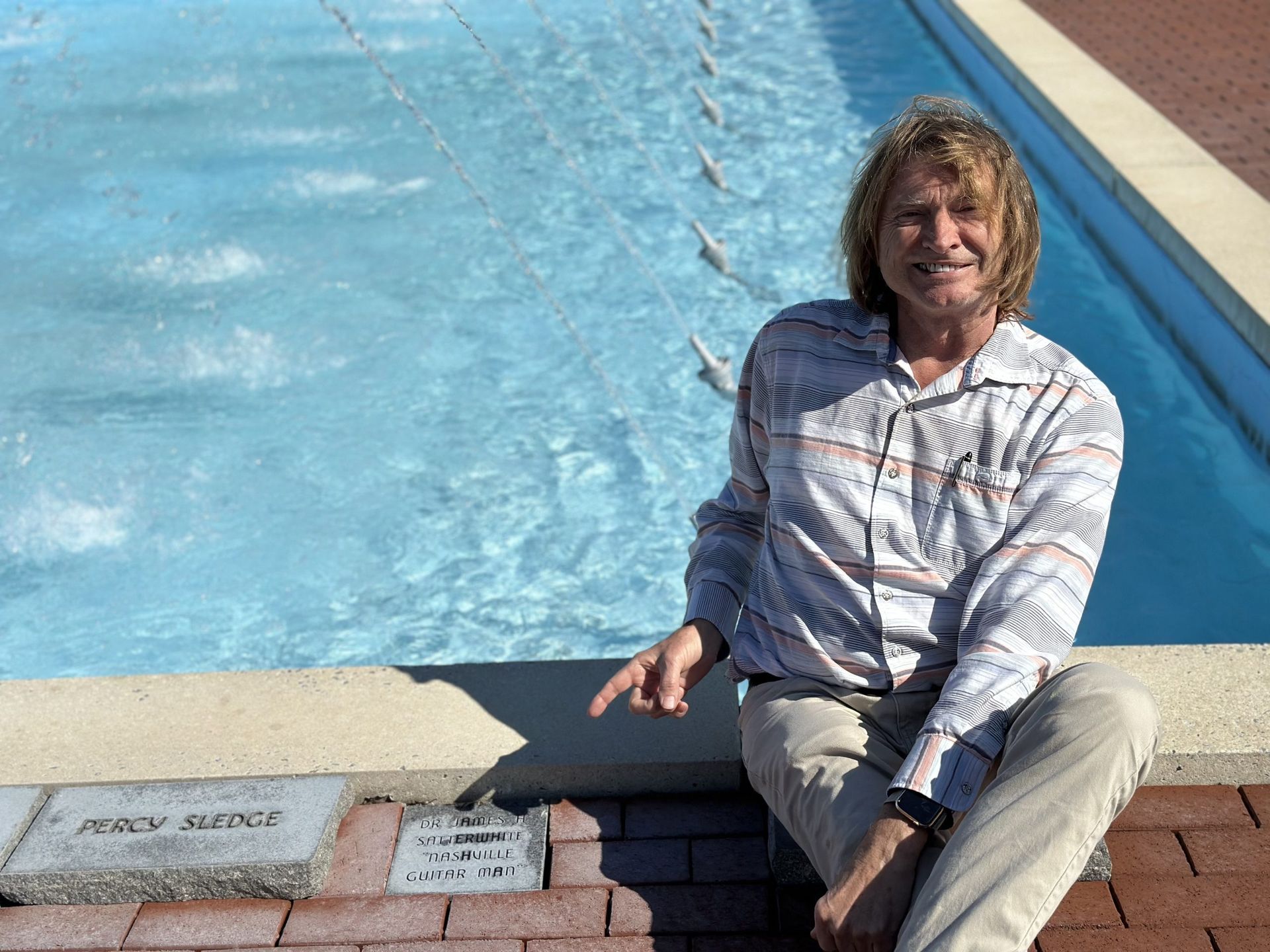 Man points at a memorial plaque next to a fountain. He sits with the fountain water behind him.