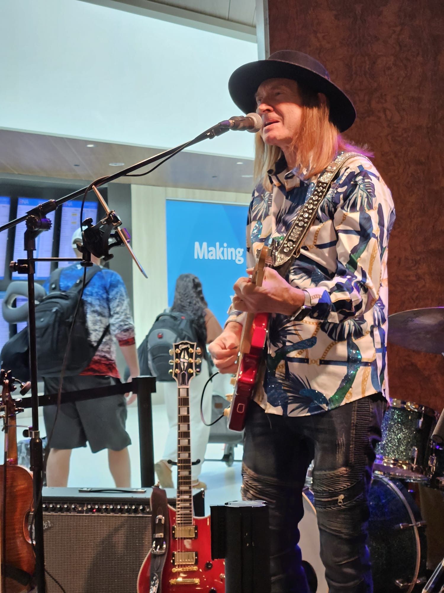 A musician with a guitar performs, wearing a hat, in an open area with other people visible.