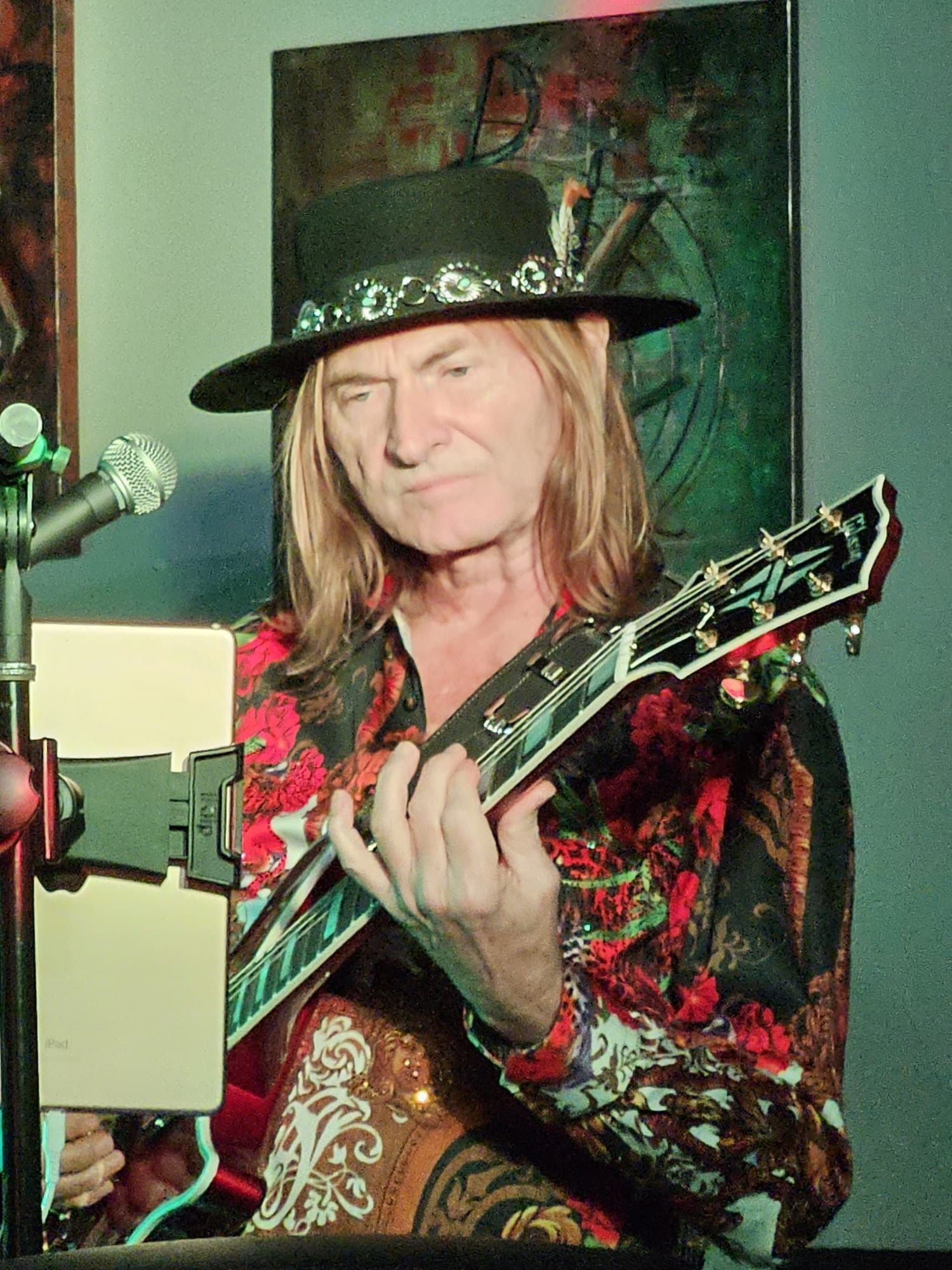 Man with long blonde hair plays guitar, wearing a decorated black hat and patterned shirt. Stage setting with microphone and screen.