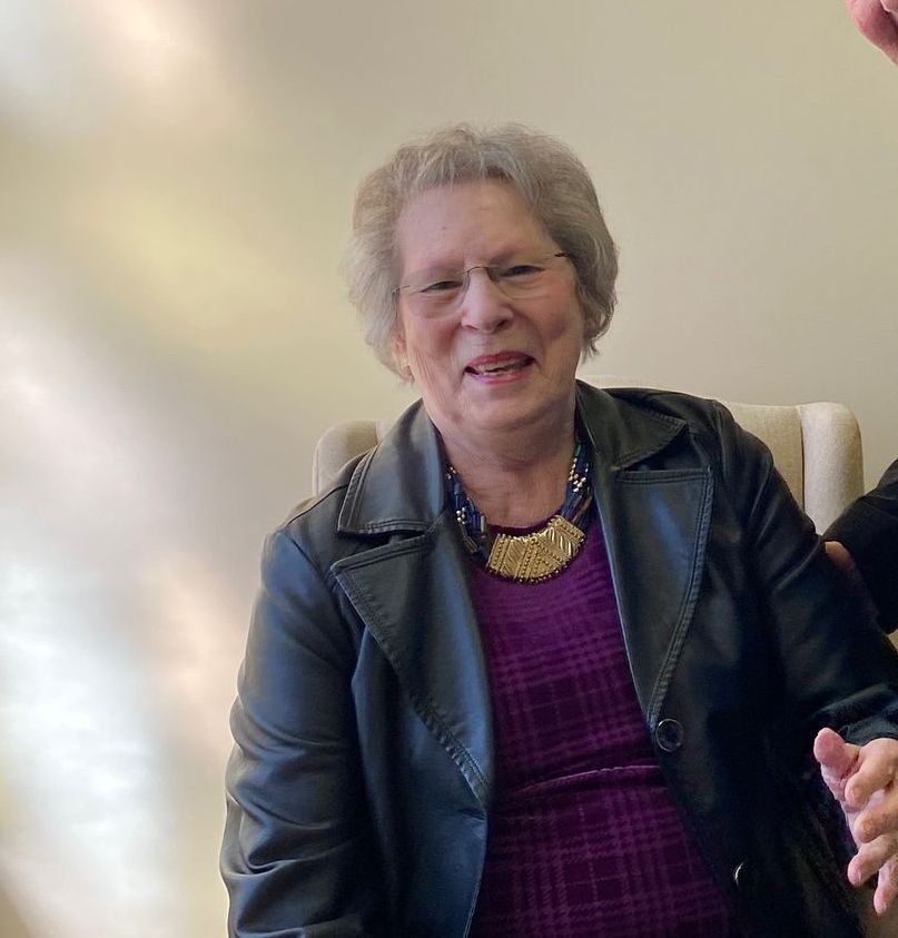 An elderly woman wearing a black leather jacket and a purple dress is sitting in a chair.