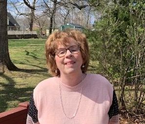 A woman wearing glasses and a pink sweater is standing in a park.