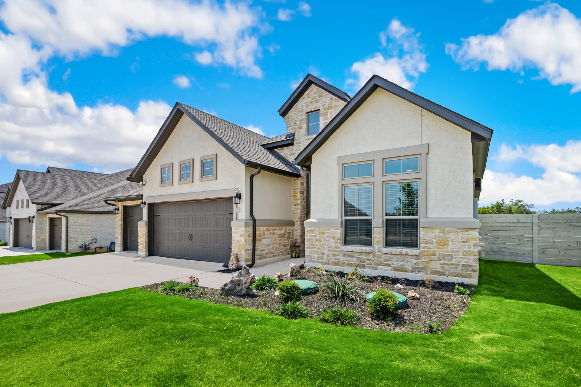 A modern, light-colored house with a stone facade, gray roof, and green lawn under a blue sky.