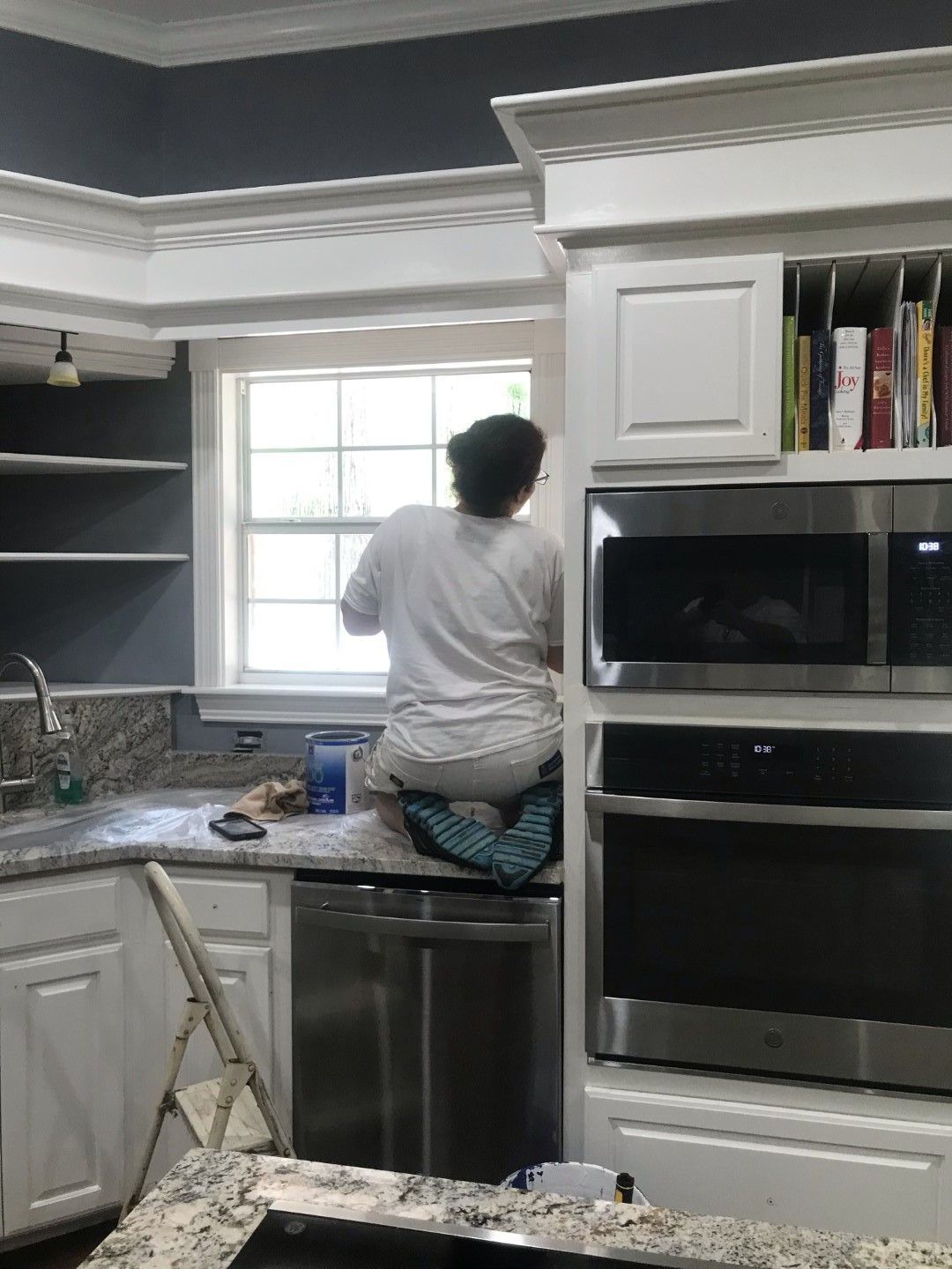 A woman is kneeling on a counter in a kitchen painting cabinets.