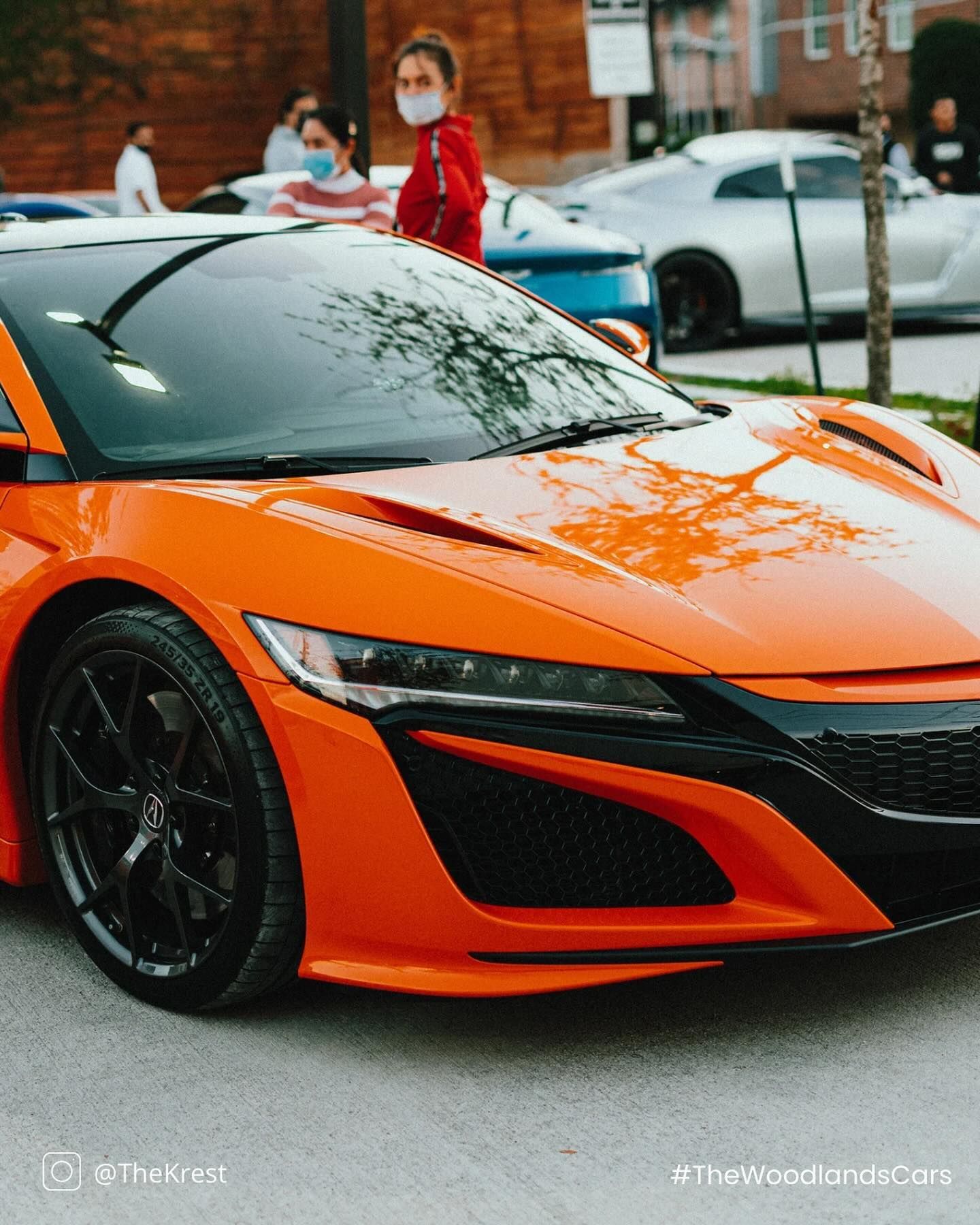 A woman wearing a mask stands next to an orange sports car