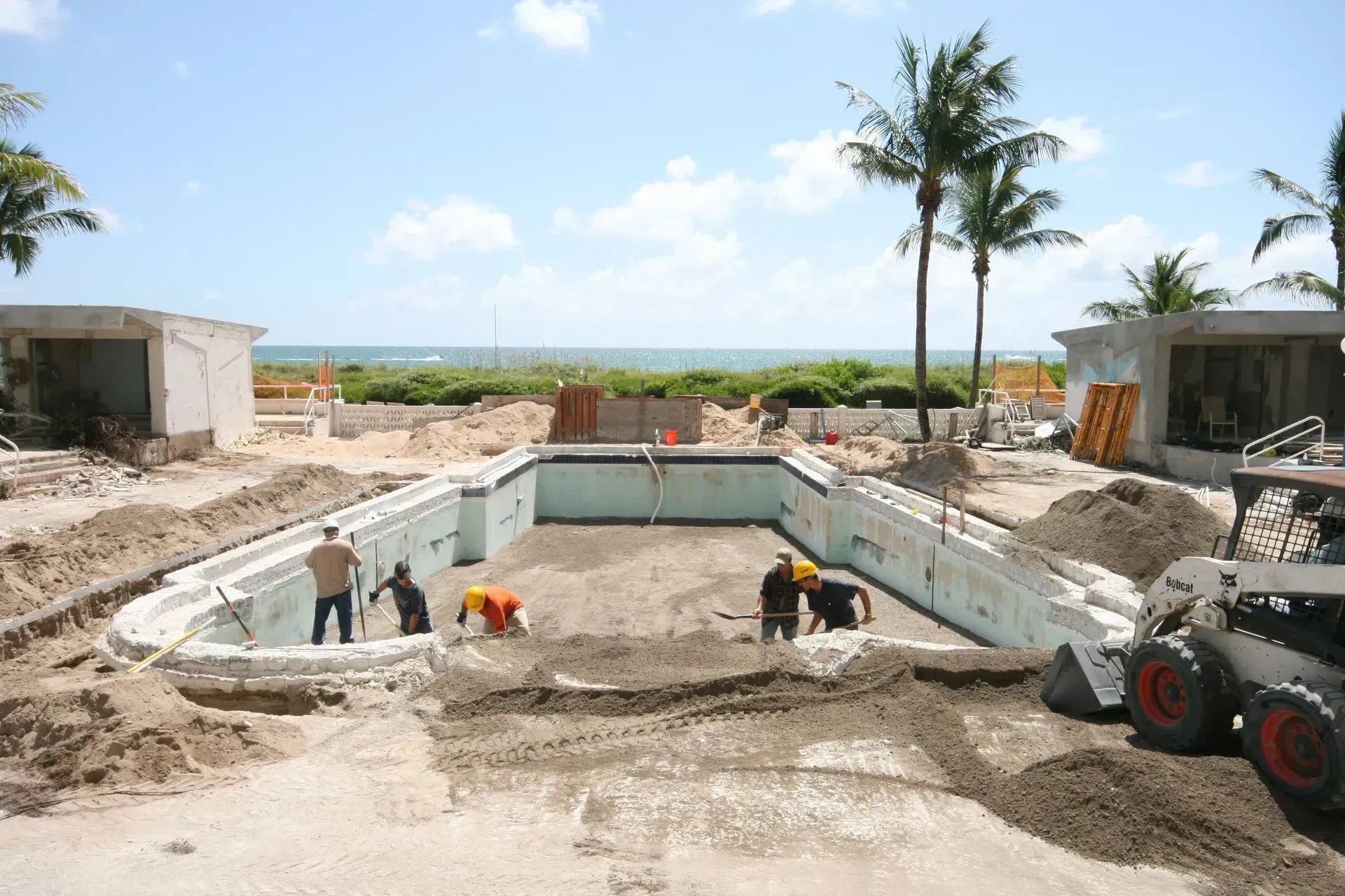 Construction site with people working in a drained pool, likely preparing to resurface.  Beach and palm trees in the background.