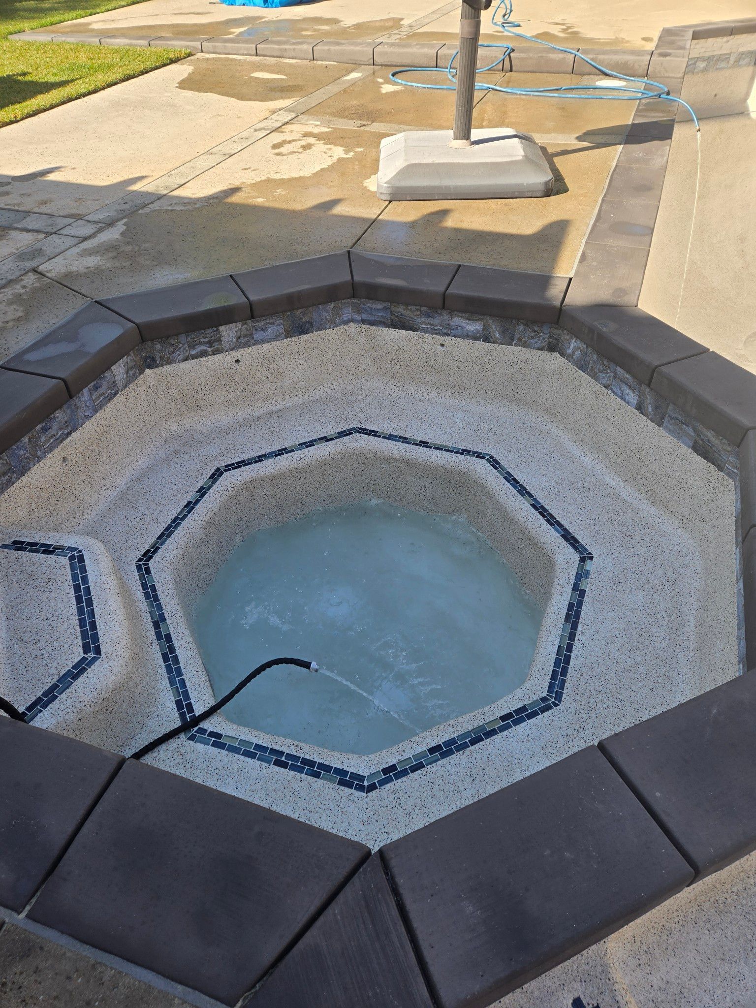 An octagonal hot tub with water, surrounded by dark gray and mosaic tile, in an outdoor setting. A black hose extends into the water.