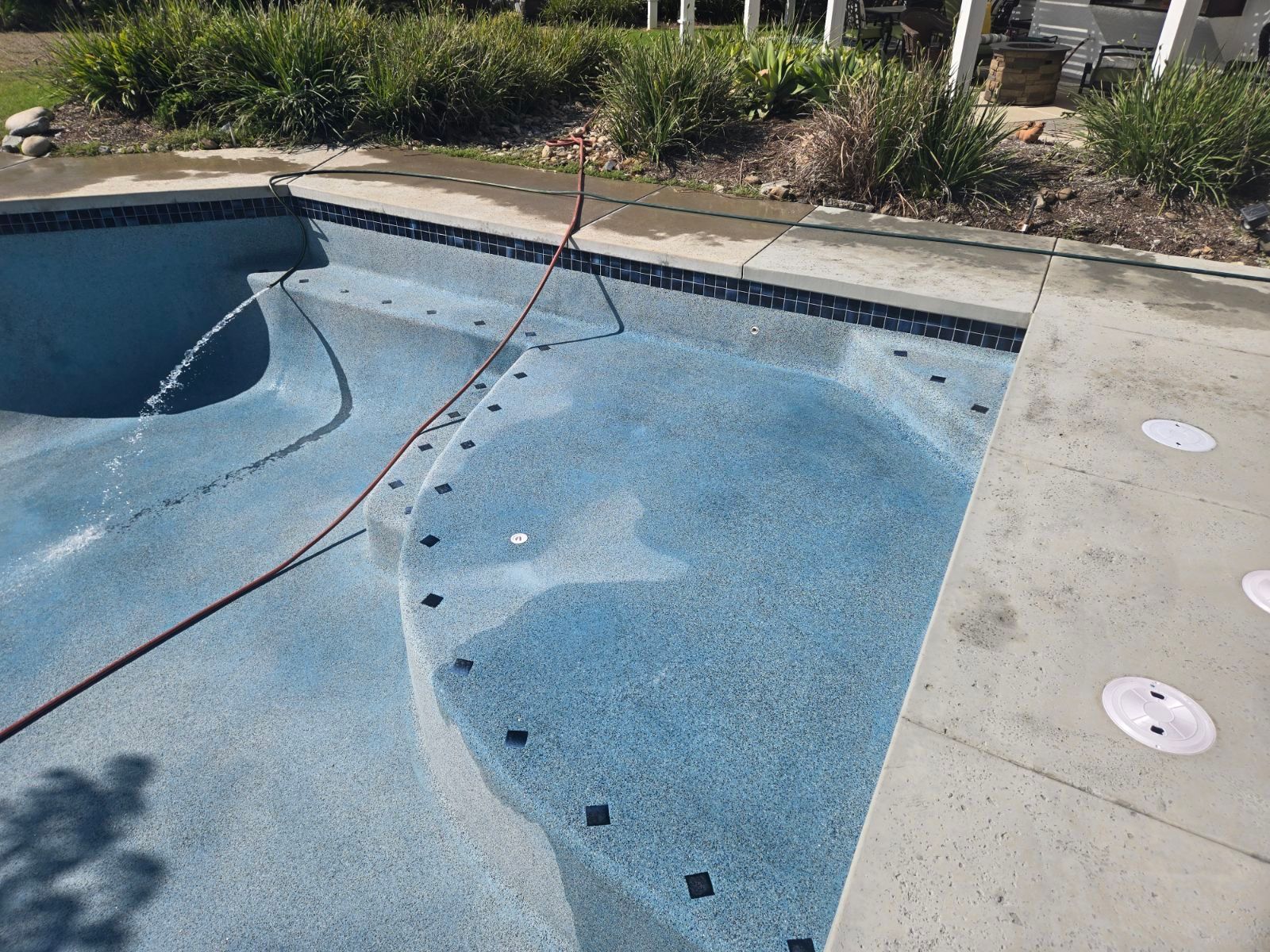 A blue-tiled swimming pool is being filled with water. A red hose lies in the pool, and plants surround the edge.