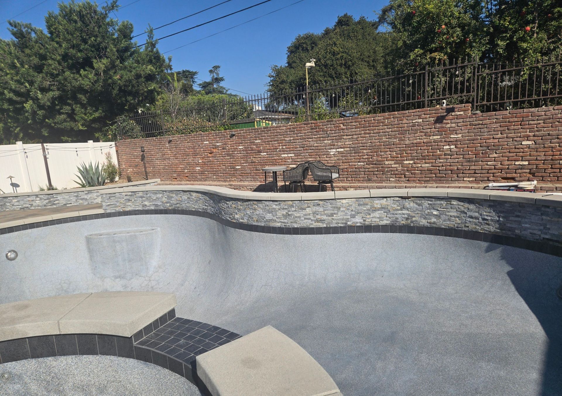 Empty swimming pool with stone coping and brick retaining wall in a sunny outdoor setting.