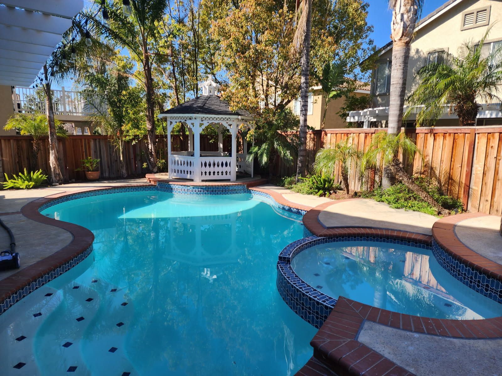 A backyard pool with a hot tub, gazebo, and wooden fence, filled with clear blue water under a bright sky.