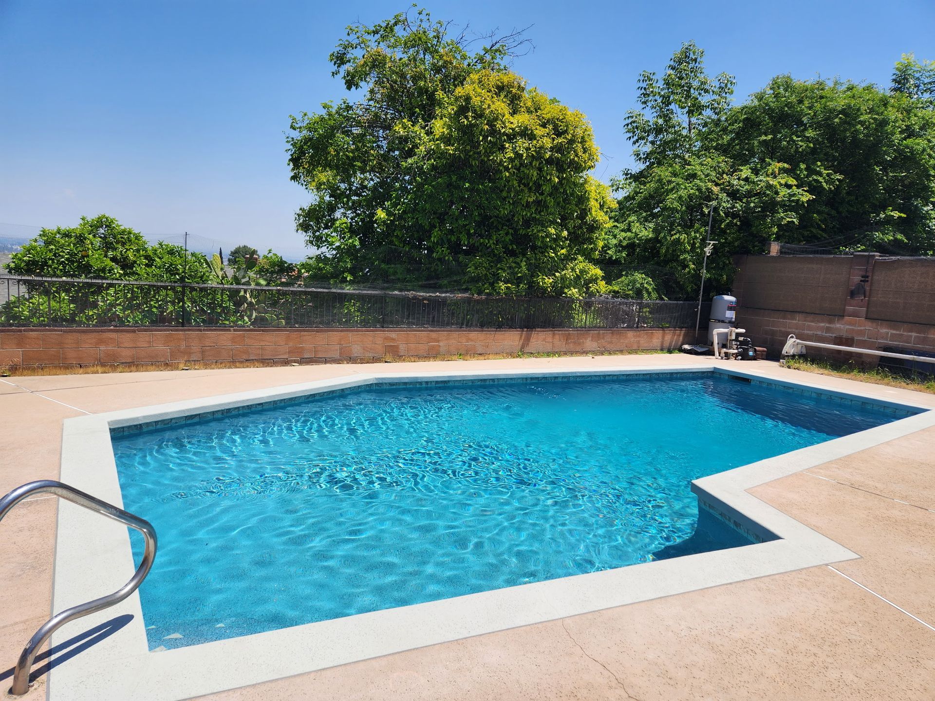 A blue swimming pool outdoors with a tan concrete deck. Lush green trees and a clear blue sky are in the background.