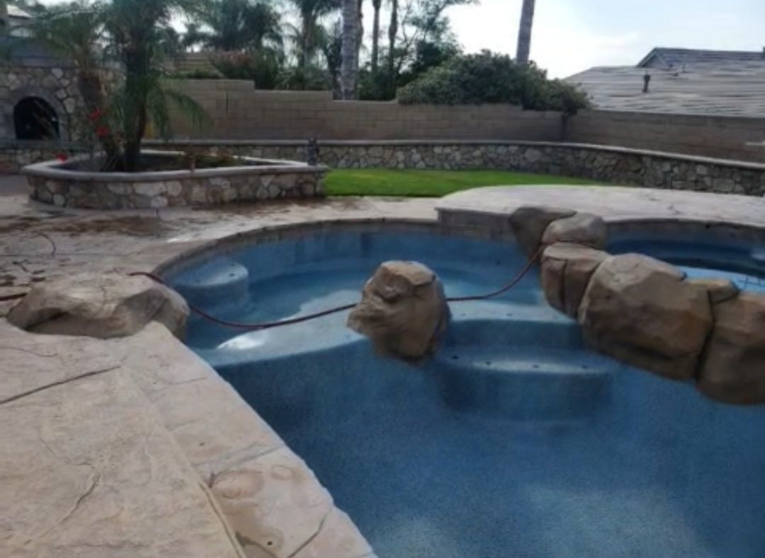 Swimming pool with rock features and a surrounding stone patio. Includes a small spa and a stone planter with a palm tree.