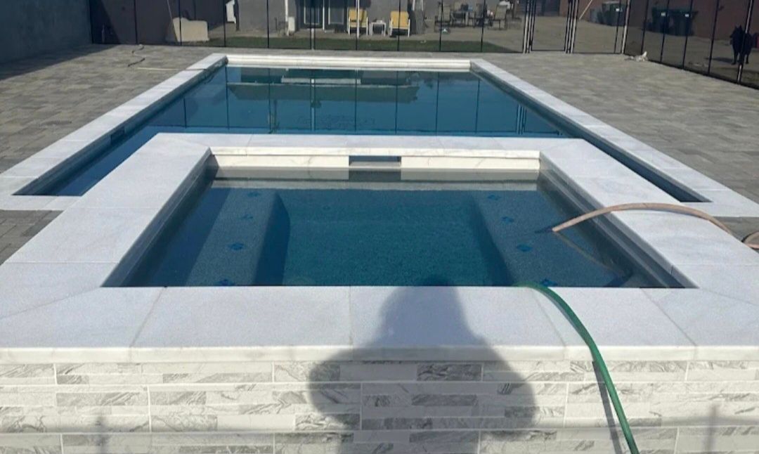 Two-tiered swimming pool with a lower, square spa section and a larger, rectangular pool above. The pool has blue-green water and white tiled borders.