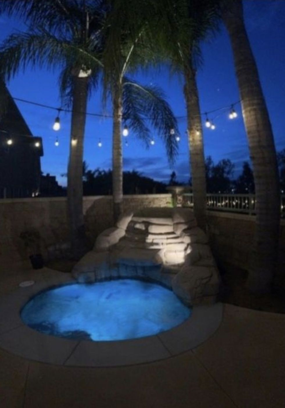 A nighttime shot of an illuminated hot tub with tiered rock features and palm trees under string lights.