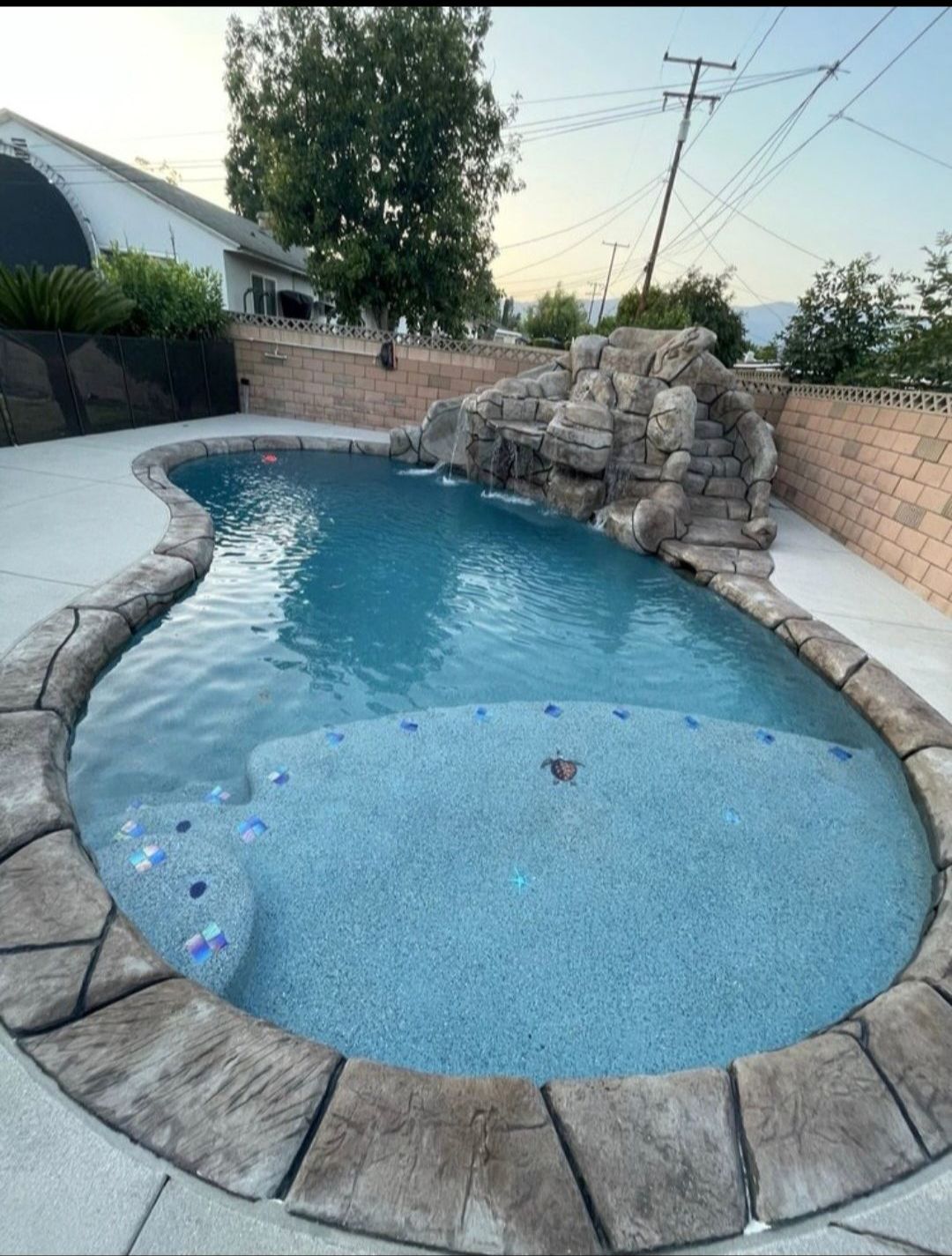 A backyard swimming pool with a waterfall feature, surrounded by a stone patio and brick wall. Blue water reflects the sky.