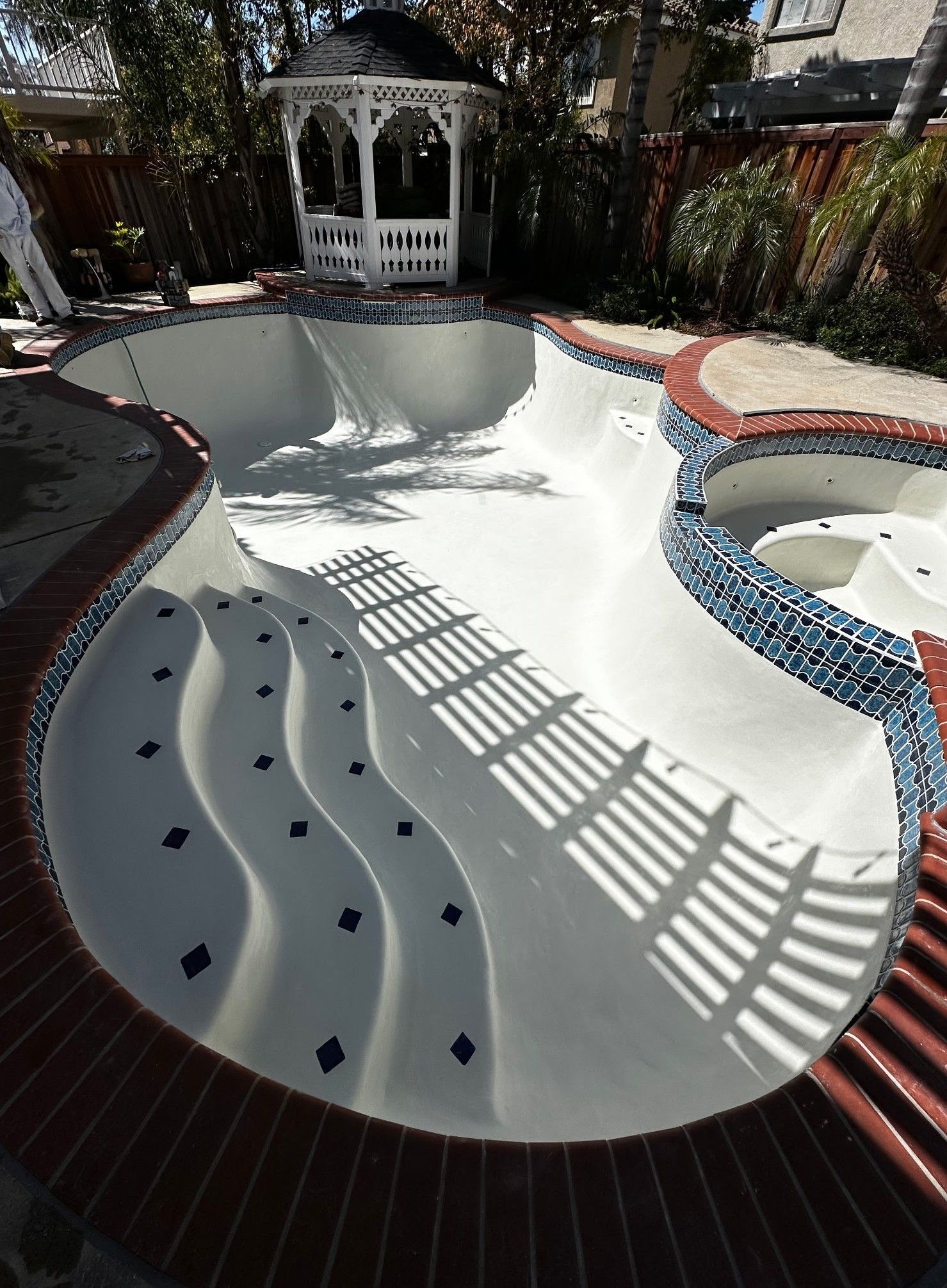 Empty, newly renovated pool with a red brick border, blue tile accents, and a white gazebo in the background.