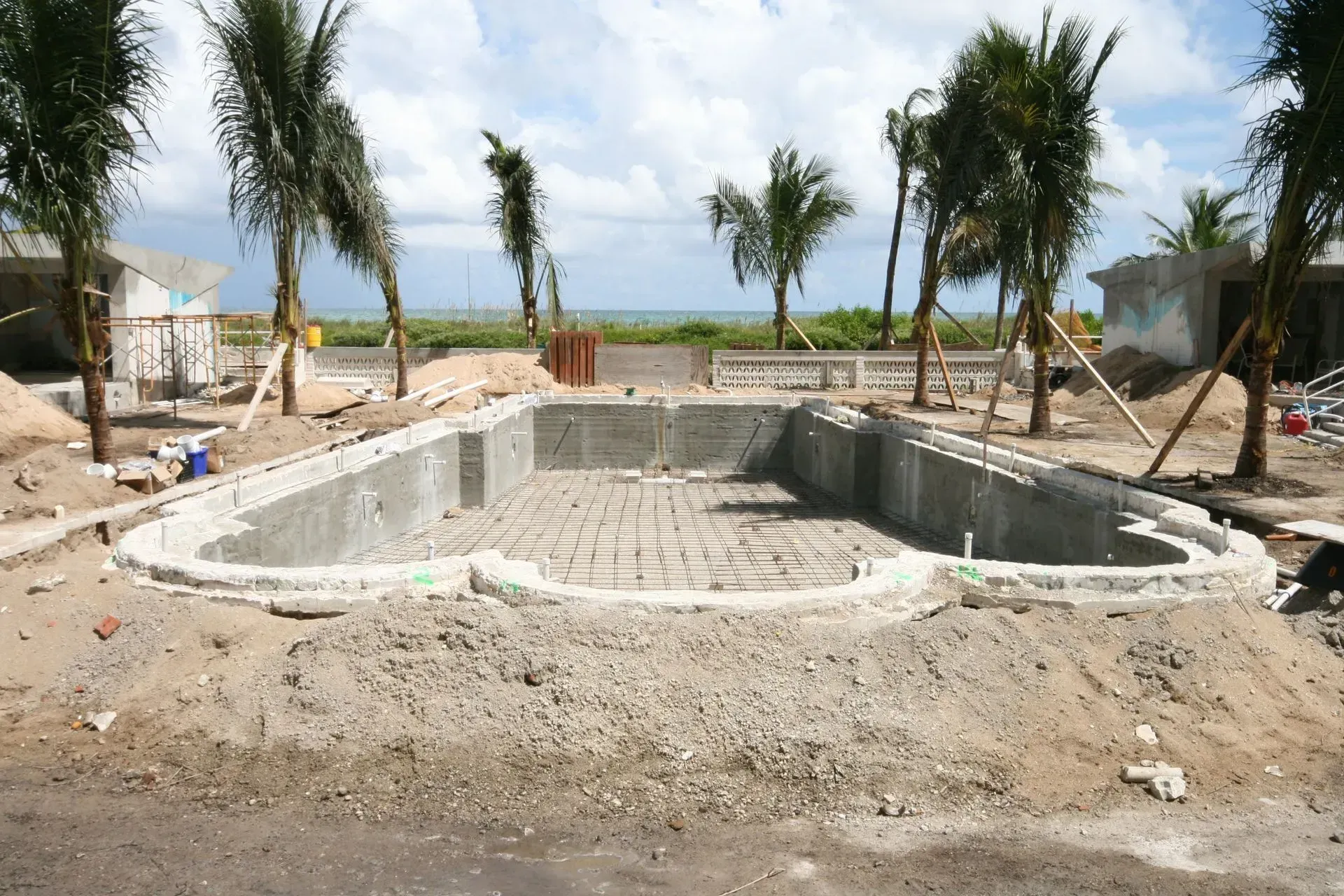A concrete swimming pool under construction outdoors with palm trees and a coastal view in the background.