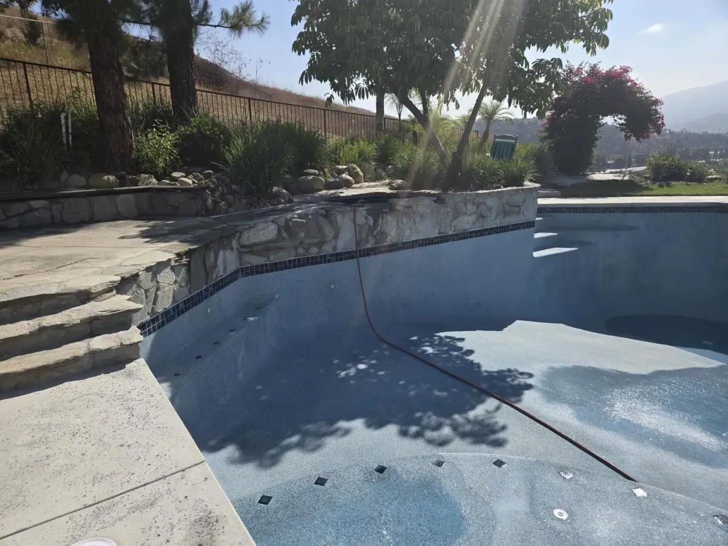 Pool with blue tiled interior, stone coping, and patio. Plants and a hillside in the background on a sunny day.