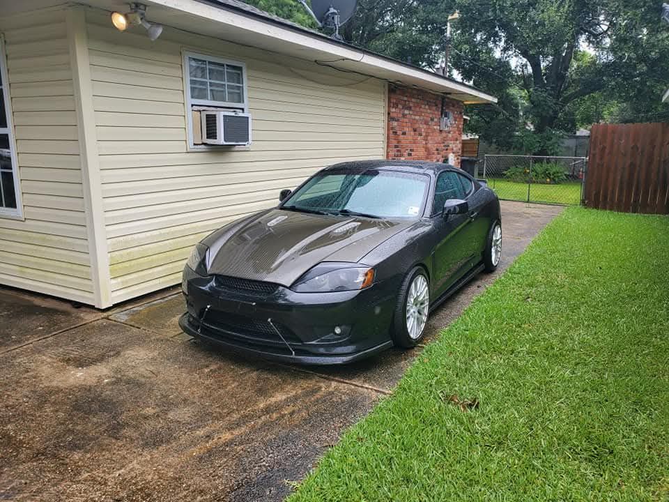 Dark gray sports car parked next to a house with beige siding, grass to the right.