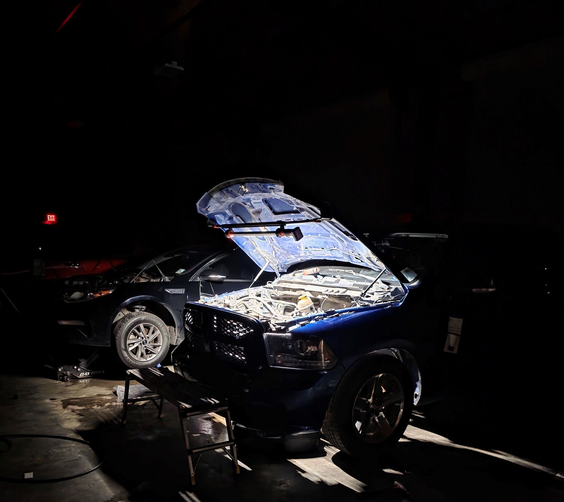 Blue car with hood open in a dimly lit garage, another car partially visible in the background.