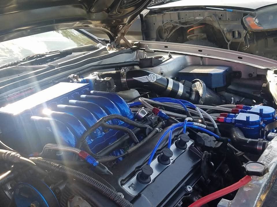Close-up of a car engine bay, showing blue intake manifold, black tubing, and various components.
