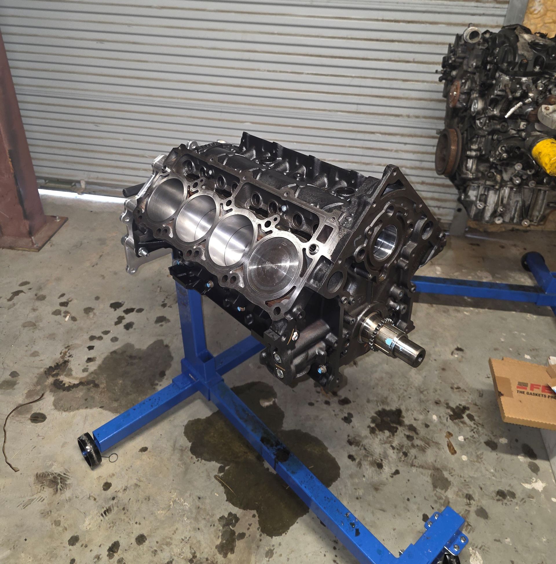 Engine block on a blue stand in a garage setting with another engine in the background.