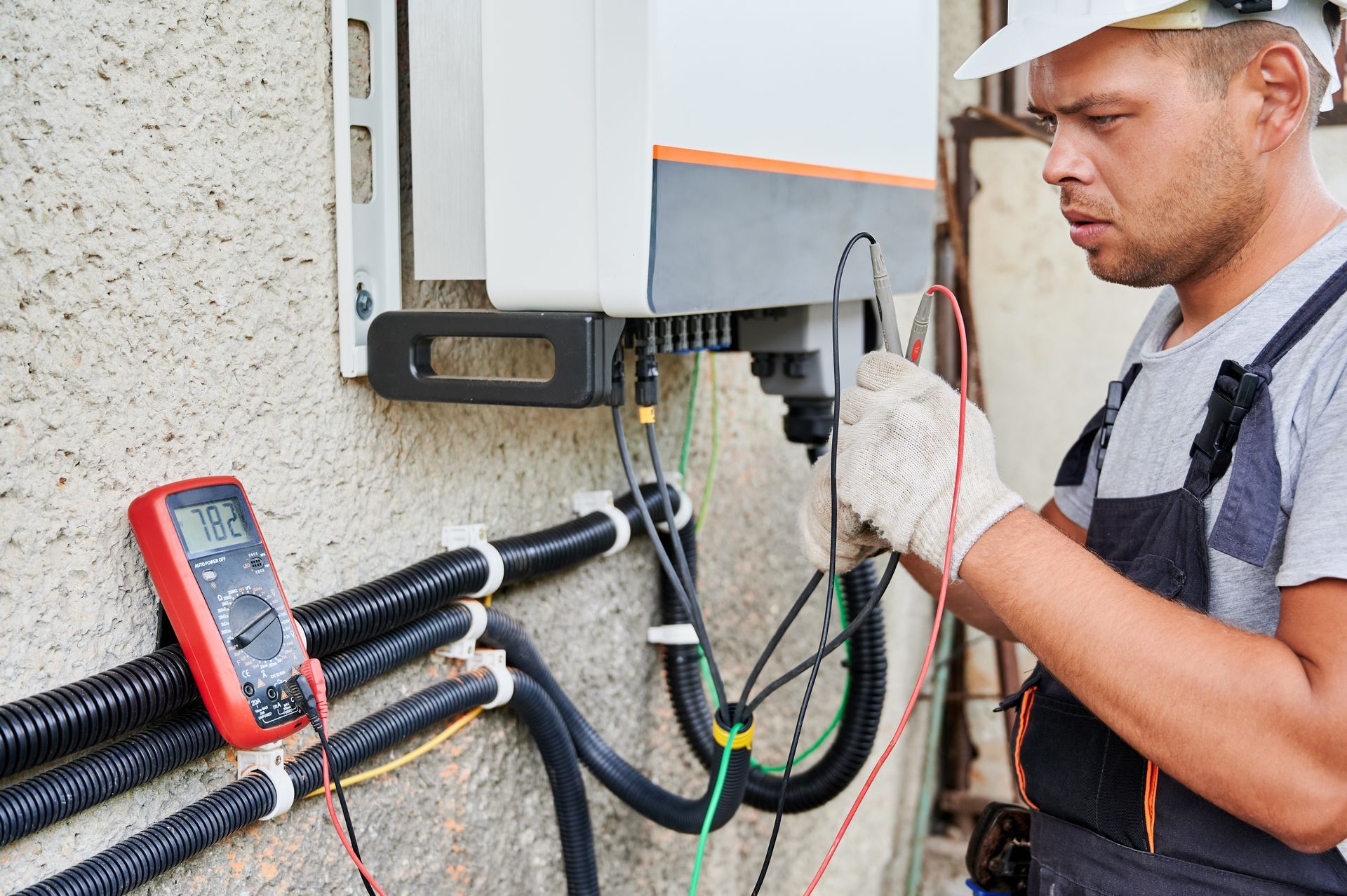 Electrician testing outdoor electrical wiring with a multimeter beside a wall-mounted unit.
