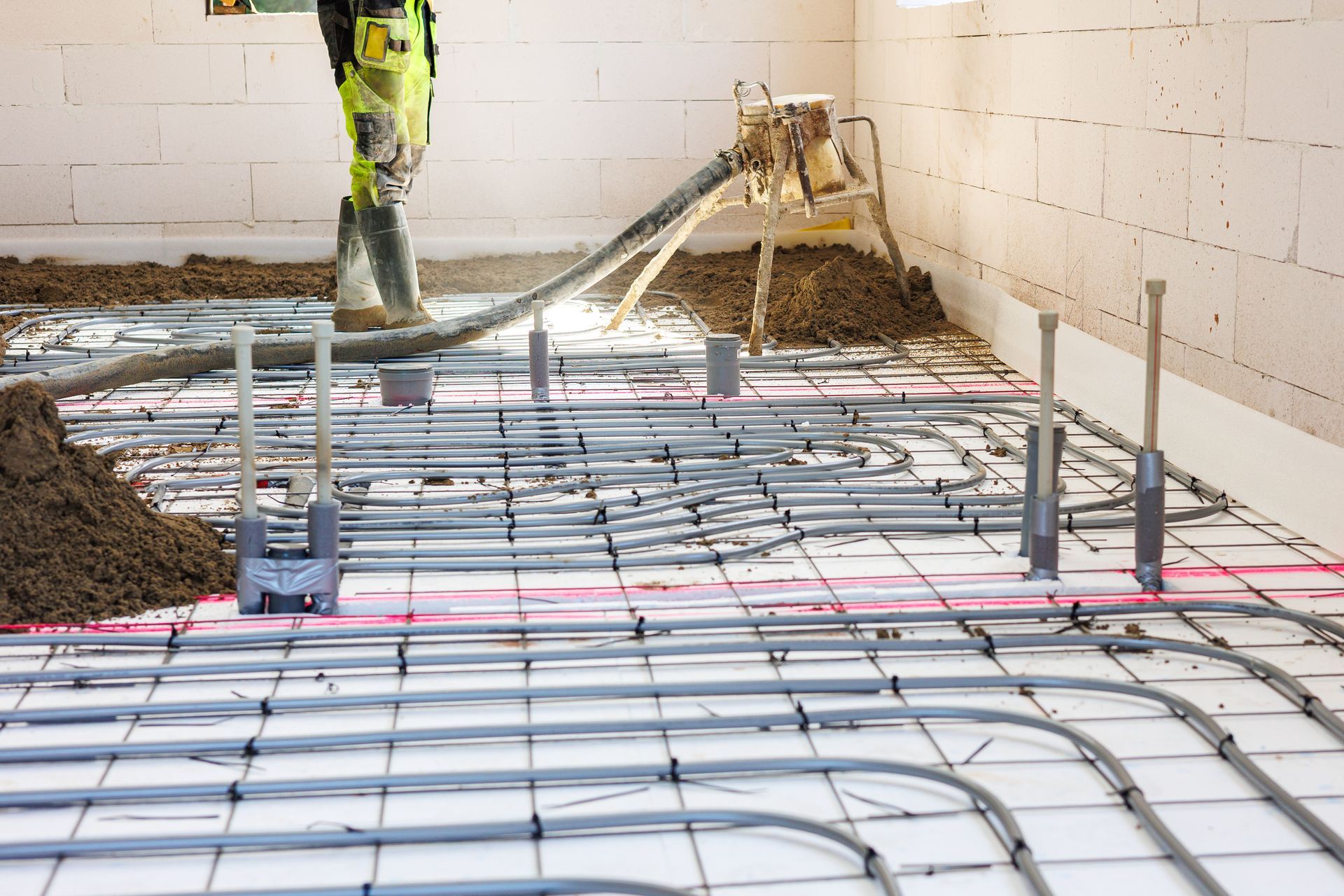 Worker pouring concrete over radiant floor heating tubes in a construction setting.