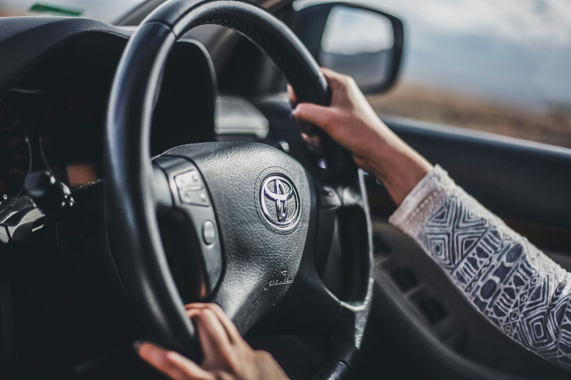 Hands on a car steering wheel with a logo, driving on a road