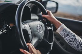 Hands on a car steering wheel with a logo, driving on a road