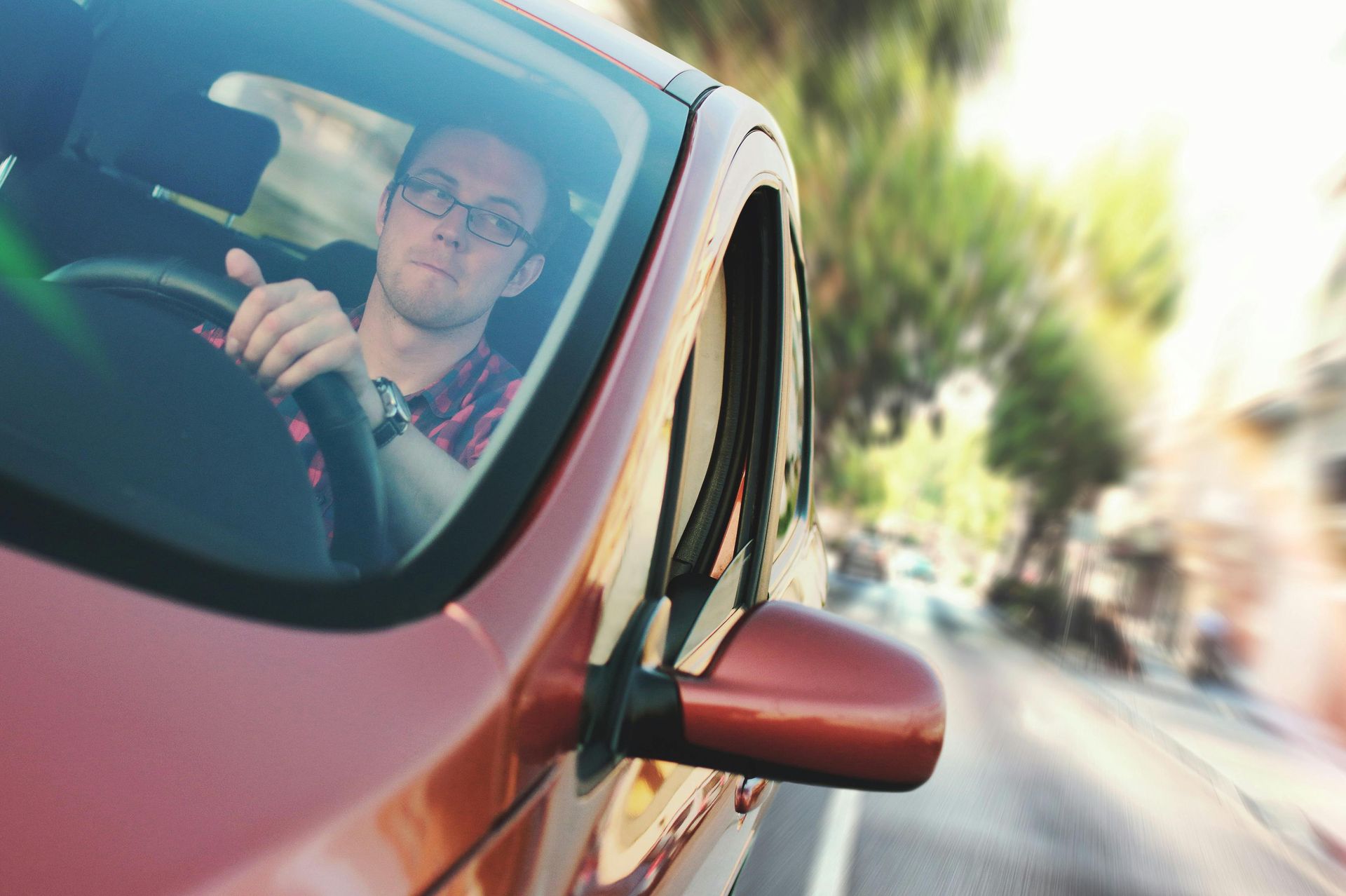 Driver seen through red car window on a blurred city street, hand on steering wheel.