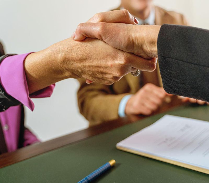 Two people shaking hands over a conference table with papers and a pen nearby