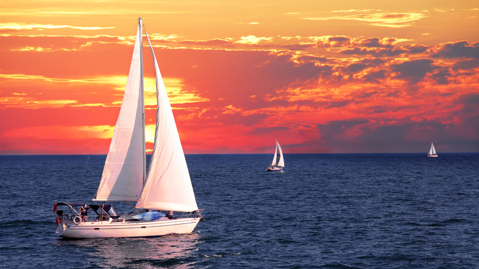 Sailboats on calm sea at sunset, with a large white sailboat in the foreground and orange sky.