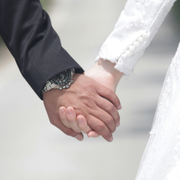 Two people holding hands, one in a dark suit and one in a white dress, with a watch visible.