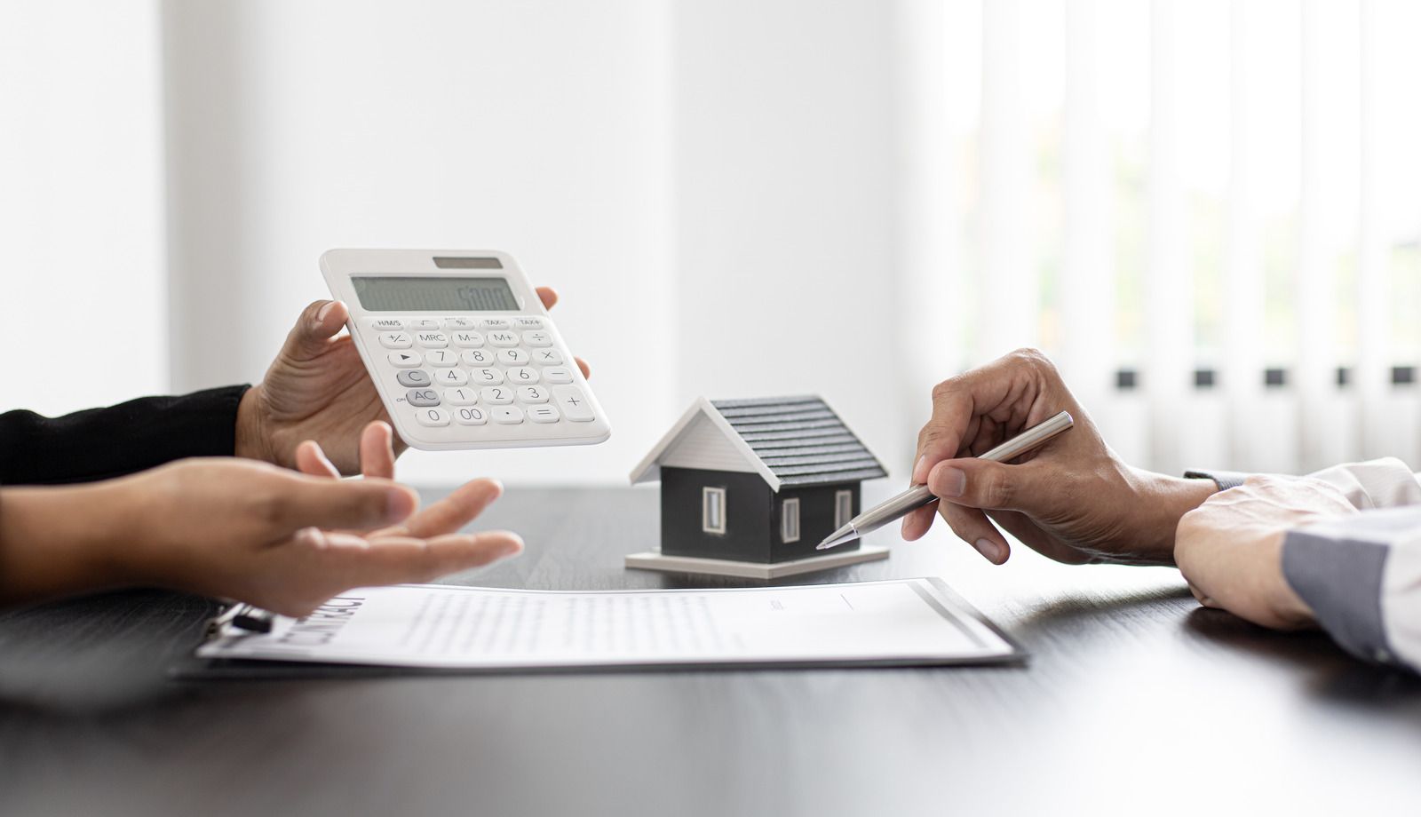 Hands calculating with calculator, signing paperwork with model house on table.