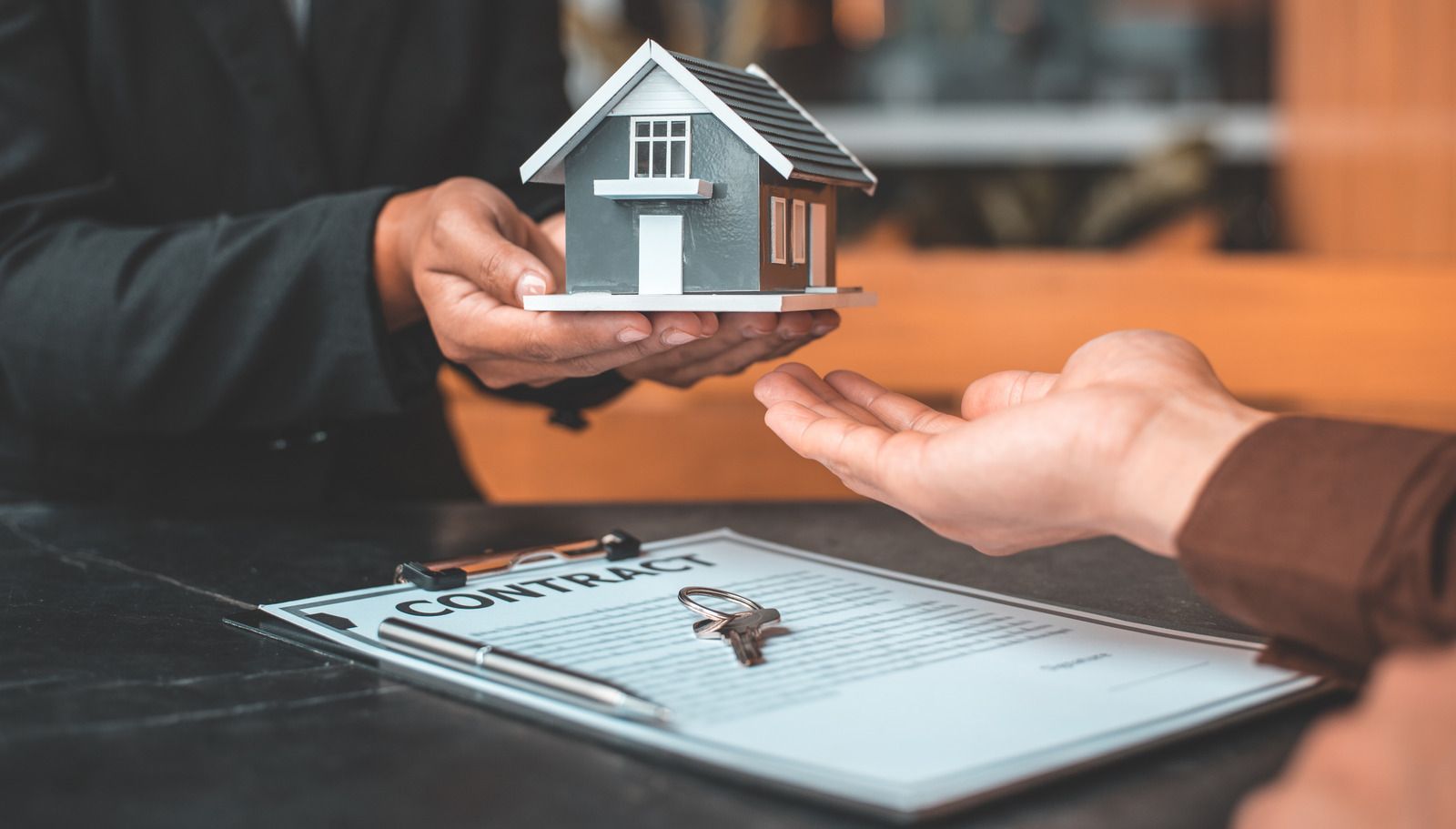 Person handing over a miniature house to another person, a contract and keys are in view.