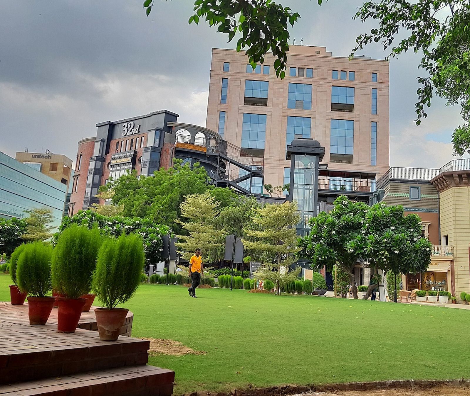 Lush green lawn with a man walking, in front of buildings, under a cloudy sky. Potted plants in the foreground.