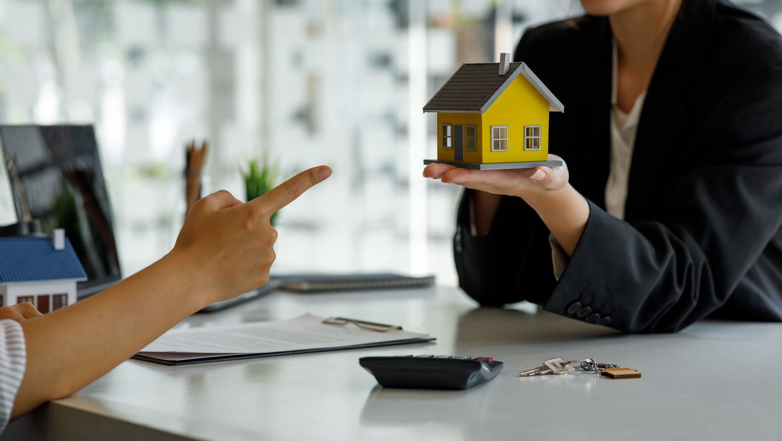 Person pointing at a miniature yellow house held by another person, likely discussing a real estate deal.