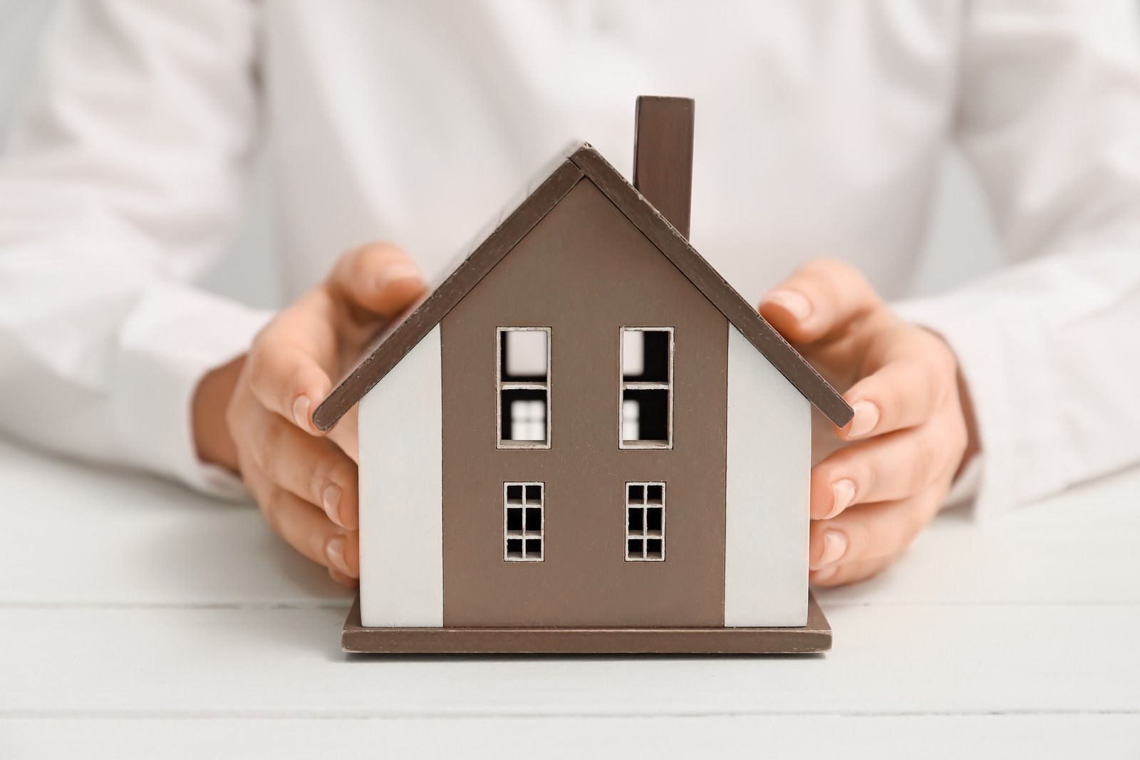 Hands holding a miniature brown house with white trim on a white surface.