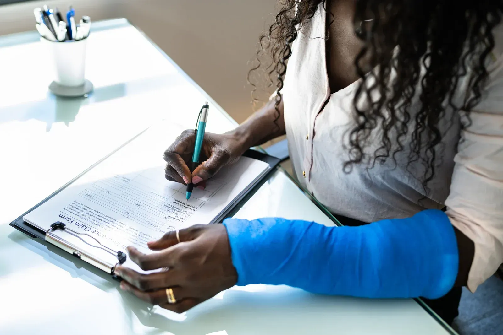 Woman with blue cast on arm signing a document at a desk, possibly completing paperwork.