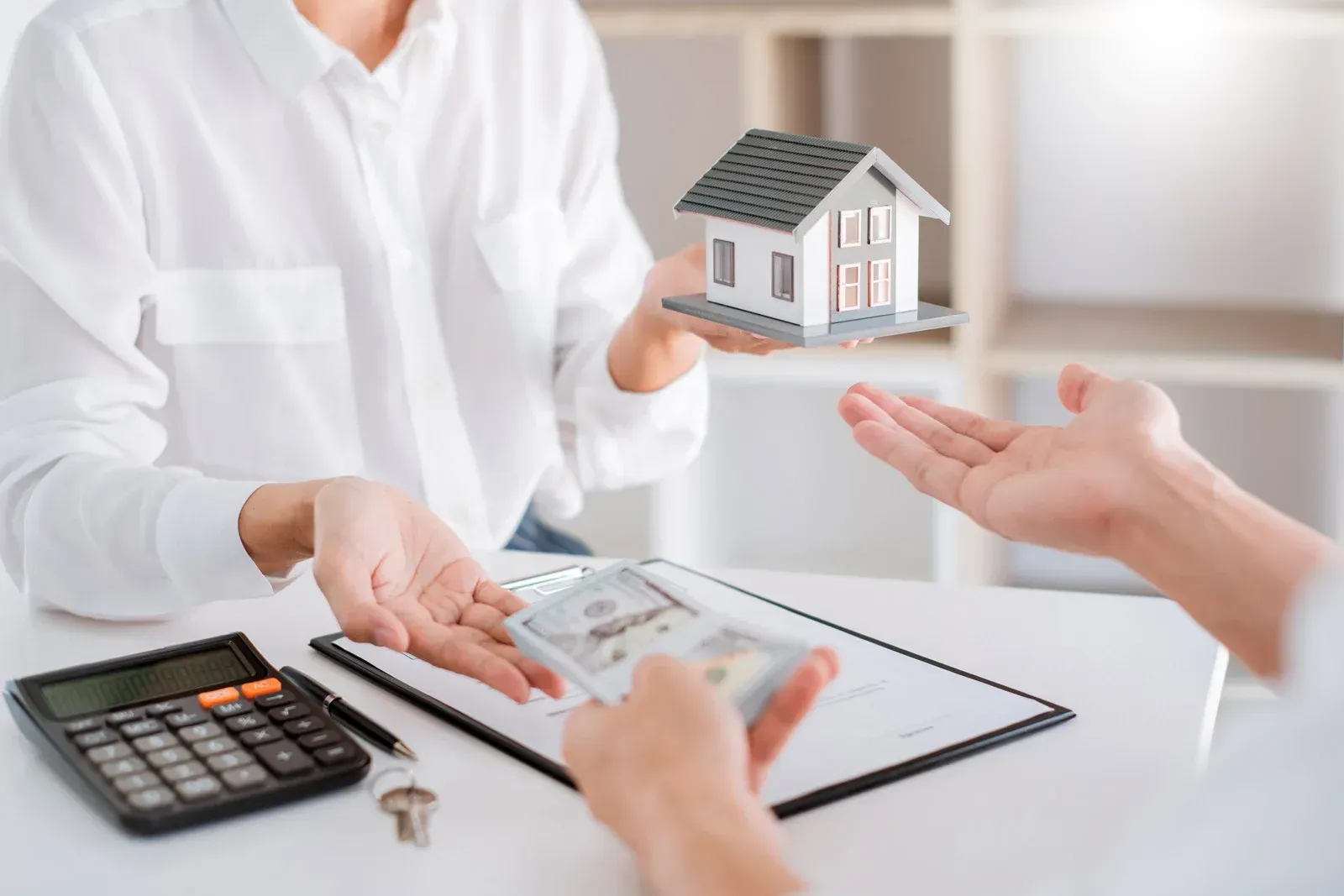 Person hands over model house, another hands cash, and keys; transaction at a table.