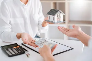 Person hands over model house, another hands cash, and keys; transaction at a table.