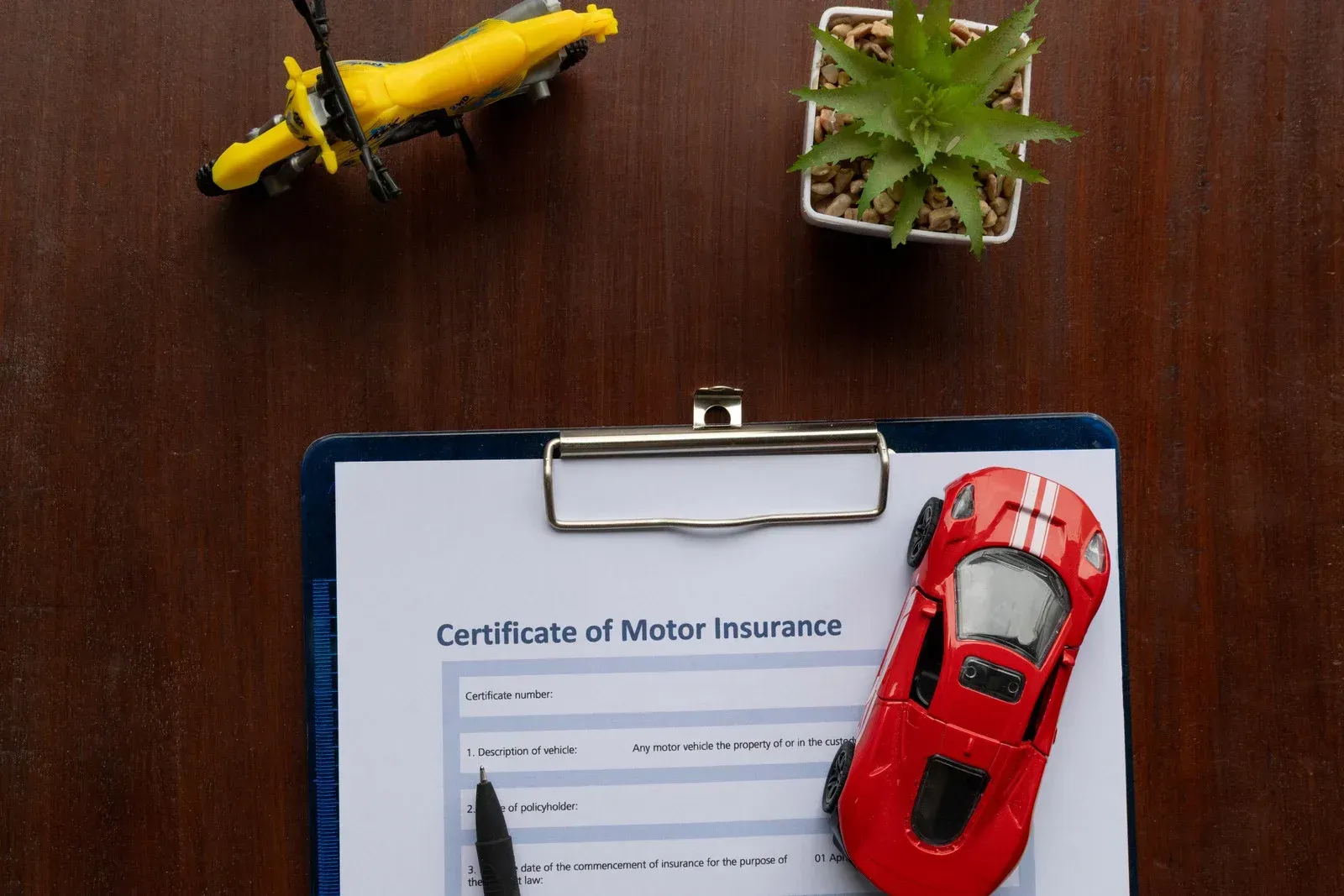 A document with a certificate of motor insurance, a toy car, a toy motorcycle, and a potted plant on a wooden surface.